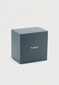 Timex-mærket kasse i mørk blå pap med en glat overflade. Forsiden prydes af et hvidt logo centreret på overfladen.