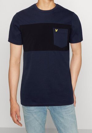 Mand iført en kortærmet t-shirt i marineblå og sort farveblok med en brystlomme, der har et lille gult fugleemblem, kombineret med lyseblå jeans.