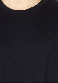 Schwarzes Baumwoll-T-Shirt mit Rundhalsausschnitt, das auf der linken Seite der Brust ein kleines schwarzes gesticktes Logo eines Vogels zeigt.