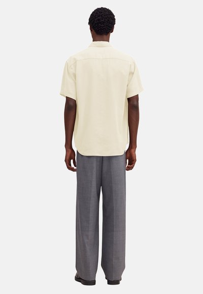Homme debout de dos, portant une chemise à manches courtes crème avec col et un pantalon gris ample, sur un fond uni.