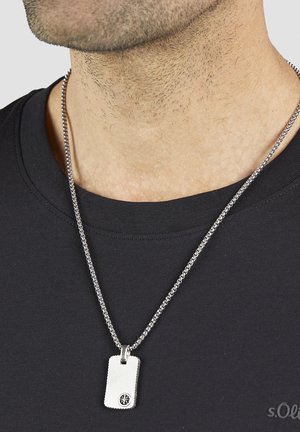 Silberne Hundemarken-Anhänger-Halskette an einer Metallkette, mit einer strukturierten Kante und einem schwarzen Emblem, getragen über einem schwarzen Hemd.