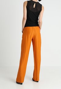 Femme debout les mains dans les poches, portant un haut sans manches noir avec un empiècement en voile et un pantalon large orange vif avec des rayures verticales.