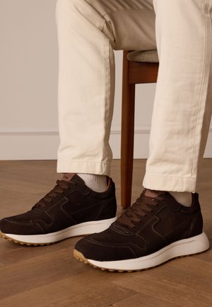 Bruine suède sneakers met een witte rubberen zool, voorzien van bruine veters, gestikt detailing en een textielen voering. Draagt op lichte broeken.