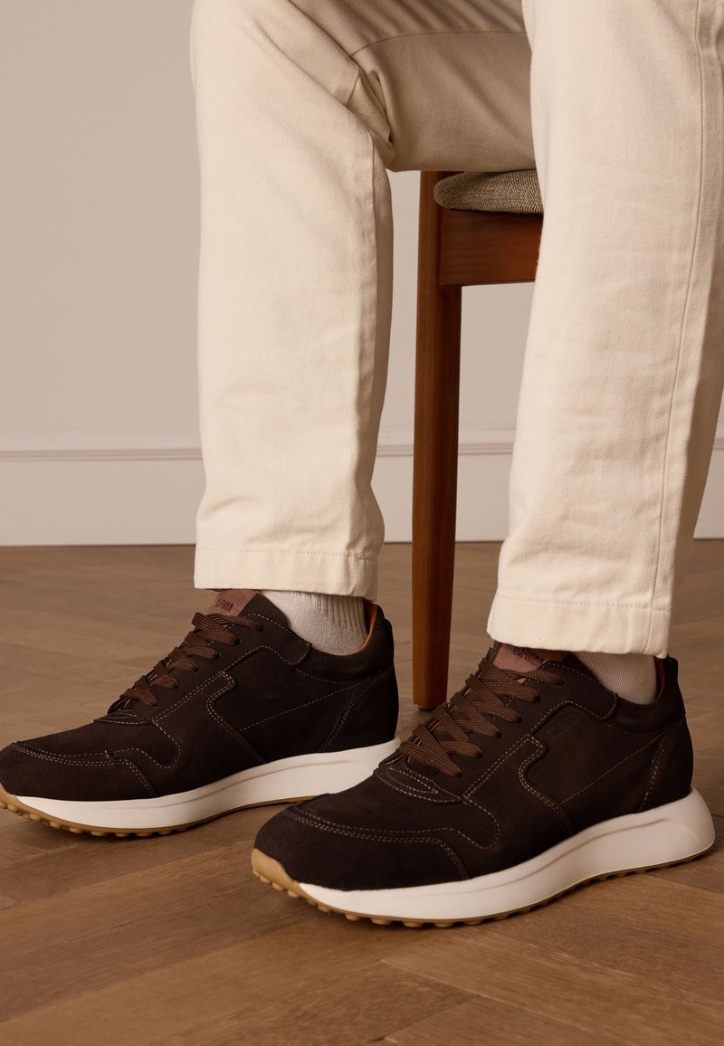 Bruine suède sneakers met een witte rubberen zool, voorzien van bruine veters, gestikt detailing en een textielen voering. Draagt op lichte broeken.
