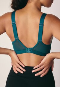 Soutien-gorge teal con spalline regolabili, dettagli in rete e chiusura con gancio e occhiello sul retro. Tessuto liscia con un design minimalista.