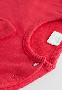 Punainen fleece-vauvan neulepaita, jossa on pyöreä kaula-aukko, ribbitetty reunus ja pieni merkkilappu. Edessä tasku ja sileä pinta.