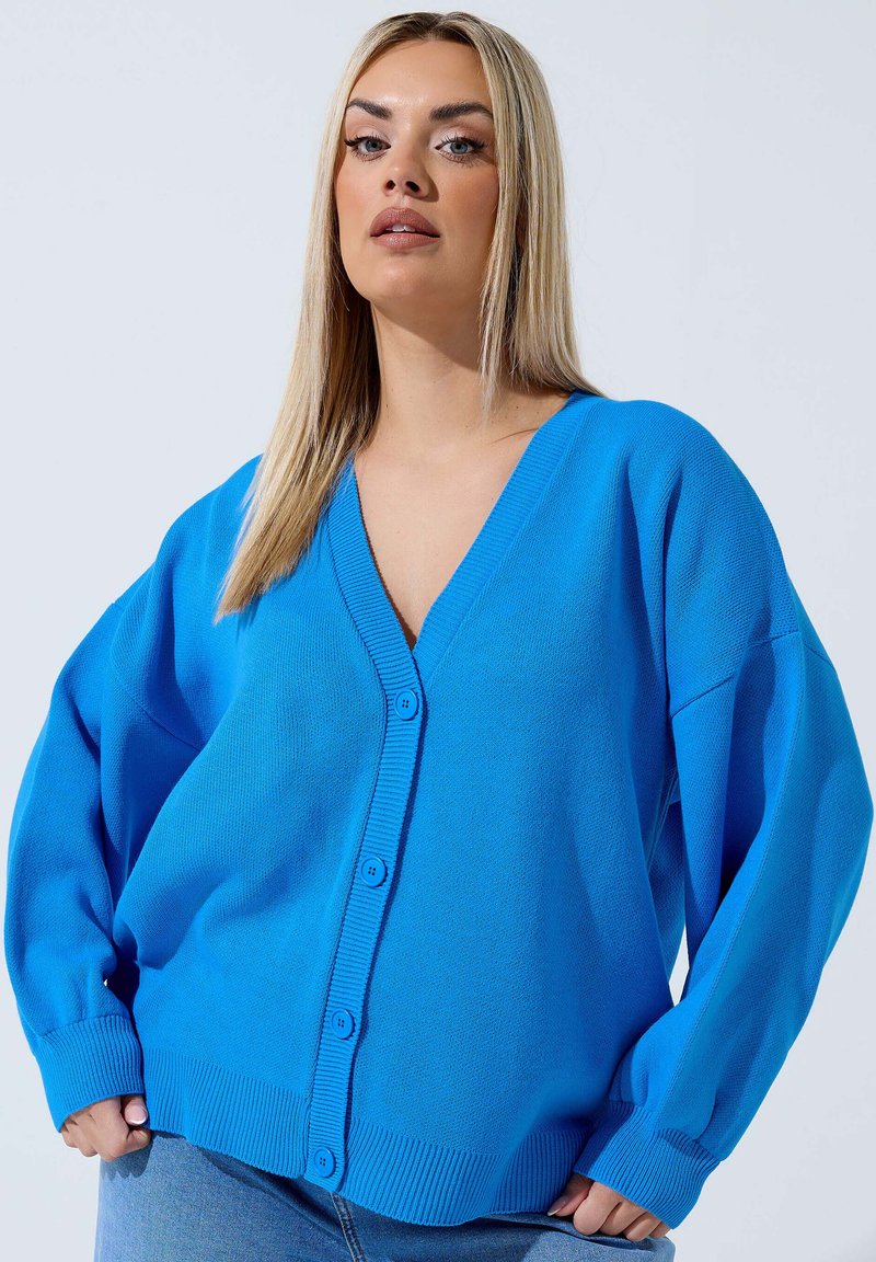 Frau mit glattem blondem Haar, die eine knallblaue, geknöpfte Strickjacke und hellblaue Jeans vor einem schlichten Hintergrund trägt.