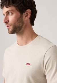 Mann mit kurzen braunen Haaren und Bart, der ein beiges Levi's-T-Shirt und eine Goldkette trägt, schaut nach links vor einem neutralen Hintergrund.