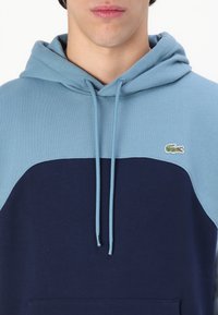 Huvtröja med färgblockdesign. Ljusblå överdel ovanför en marinblå underdel. Har en liten broderad logotyp.