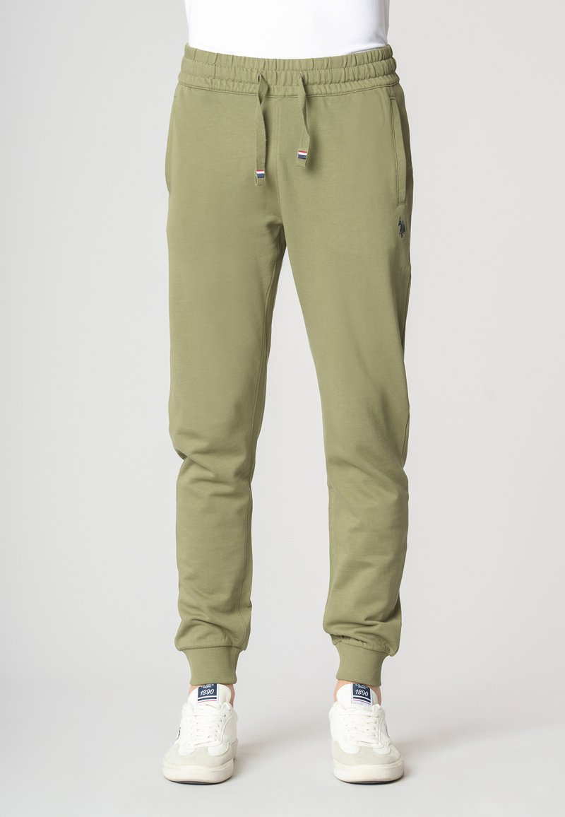 Pantaloni jogger verde oliva con vita elastica, cordoncino, tasche laterali e polsini a costine. Abbinati a sneakers bianche e grigie.