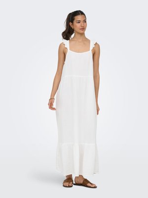 Jonge vrouw met donker haar in een paardenstaart, draagt een witte mouwloze maxi-jurk met gerafelde bandjes en bruine open-toe sandalen, staand.