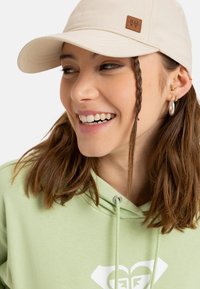 Gorra de béisbol beige con un parche de logo de cuero marrón, combinada con una sudadera verde claro que presenta un diseño gráfico blanco.