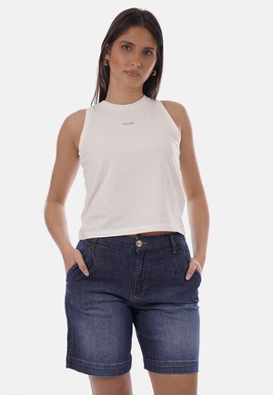 Giovane donna con lunghi capelli scuri che indossa una canotta bianca senza maniche e pantaloncini di jeans blu con le mani nelle tasche, sfondo neutro.