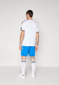 Hvid sportstrøje med sorte skulderdetaljer, parret med blå shorts. Hvide strømper med sorte striber og hvide fodboldstøvler.
