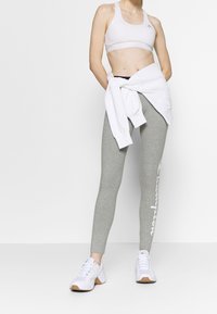 Szare legginsy z białymi detalami logo, w połączeniu z białym stanikem sportowym oraz przewiązaną w talii białą bluzą. Całość dopełniają białe buty sportowe.