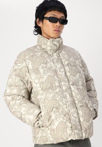 Padded jacket with a high collar, featuring a beige paisley pattern, elastic cuffs, and a front zip closure. Textured fabric.