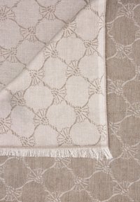 Beżowa tkana tkanina z misternym wzorem okręgów i akcentami przypominającymi kokardki. Krawędzie zdobione delikatnym frędzlem.