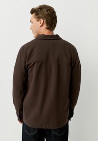 Camisa marrón con cuello, hecha de una tela suave, que presenta una costura recta en la parte trasera y mangas largas, combinada con pantalones vaqueros oscuros.