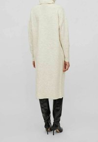 Abito a maglia color crema con collo alto, maniche lunghe, lunghezza al ginocchio, dal taglio morbido e con una leggera texture; abbinato a stivali neri lucidi alti fino alla coscia.