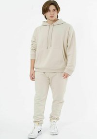 Beiger Hoodie mit einer Vordertasche und Kordelzug, kombiniert mit passenden tapered Sweatpants. Getragen mit weißen High-Top Sneakers.