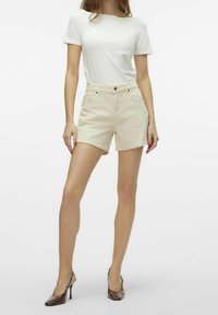 Pantalones cortos de mezclilla beige con cintura alta, bolsillos frontales y dobladillos deshilachados, combinados con una camiseta básica blanca de manga corta y tacones altos marrones.