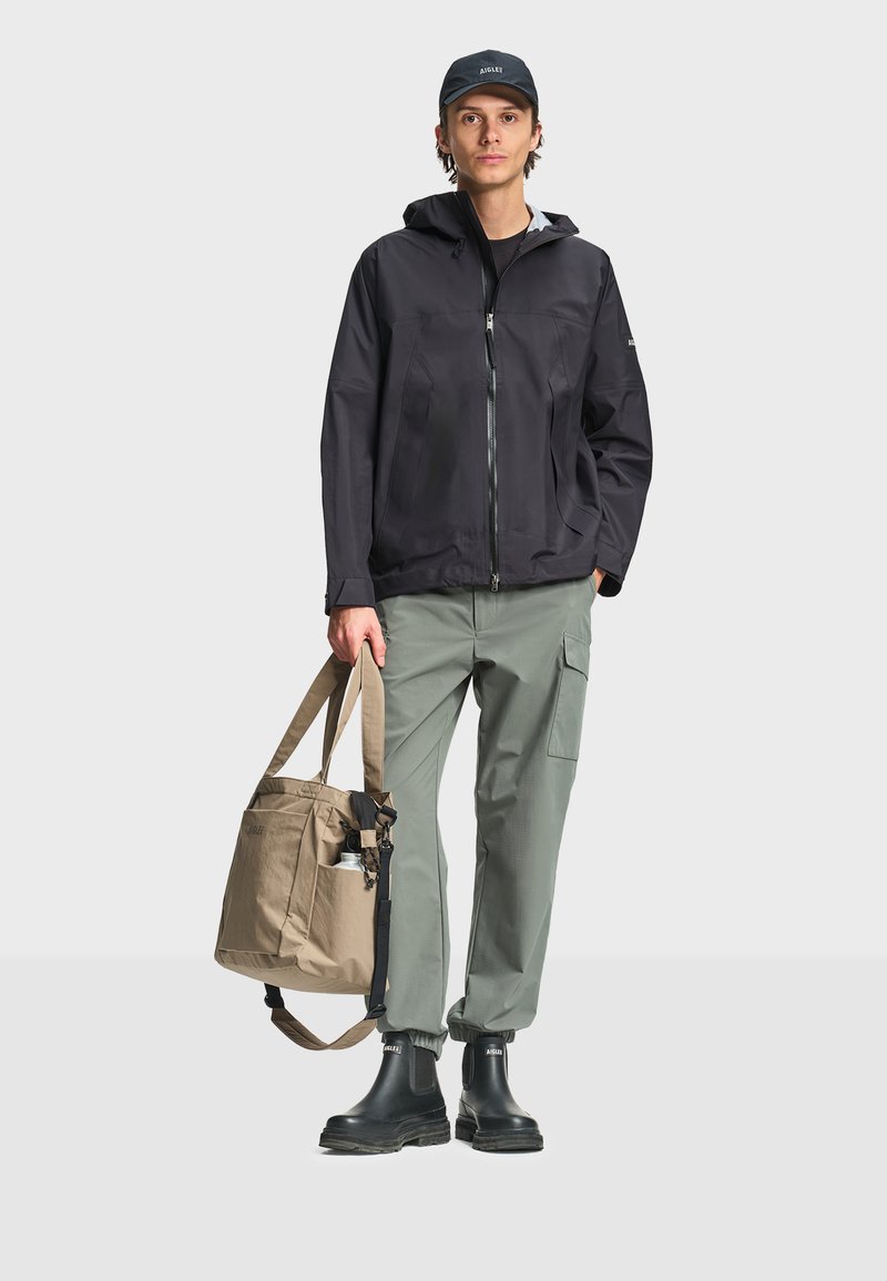 Jeune homme portant une veste à capuche noire, un pantalon cargo vert, des bottes noires et une casquette, tenant un sac fourre-tout beige avec une bouteille d'eau dans la poche latérale.