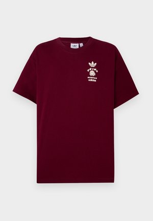 Mężczyzna ubrany w bordową koszulkę Adidas z białym logo i japońskim napisem, czarne spodnie z białymi paskami po bokach oraz z tatuażem na prałym przedramieniu.