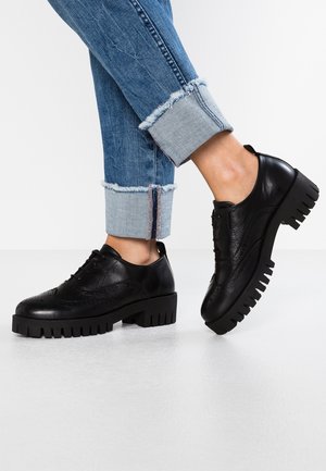 Sorte brogue-sko med chunky sål, brugt med opkrogede blå denimjeans på hvid gulvbaggrund.