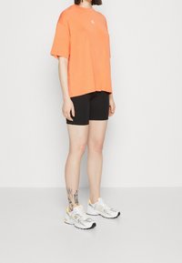 Camiseta naranja oversized con cuello redondo, pantalones cortos negros ajustados y zapatillas blancas con acentos amarillos y patrones detallados.