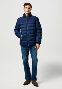 Mørkeblå pufferjakke med høj krave, lynlåslukning, vandret quiltning, to frontlommer og en struktureret design på skuldrene.