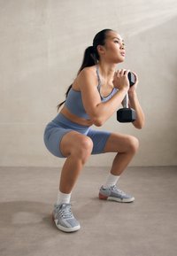 Vrouwelijke atleet in een lichtblauwe crop top en shorts, die een squat uitvoert terwijl ze een zwart dumbbell vasthoudt. Ze draagt grijze sportschoenen met roze accenten.