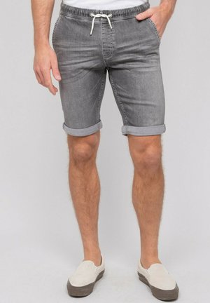 Personne portant un short en jean gris retroussé à taille à cordon, des chaussures légères à enfiler et un t-shirt blanc, debout devant un fond uni.