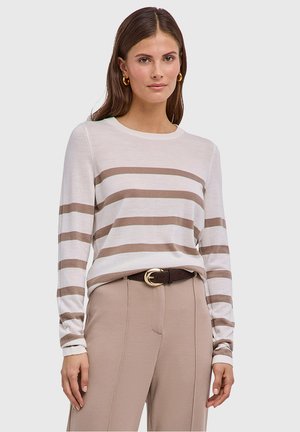 Frau trägt ein langärmliges Top mit beigen und weißen Streifen, beigefarbene Hose mit braunem Gürtel und goldene Creolen, steht vor einem einfarbigen Hintergrund.