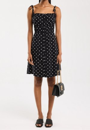 Nainen, joka pitää mustaa, valkoisilla pilkuilla koristeltua hihattomaa mekkoa, mustaa käsilaukkua, jossa on kultaiset ketjuolkaimet, ja mustia kudottuja liukusandaaleja.