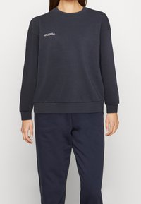 Felpa blu navy in morbido tessuto, con scollatura a girocollo, maniche lunghe e una piccola stampa di testo bianca sul petto. Polsini e orlo texture.