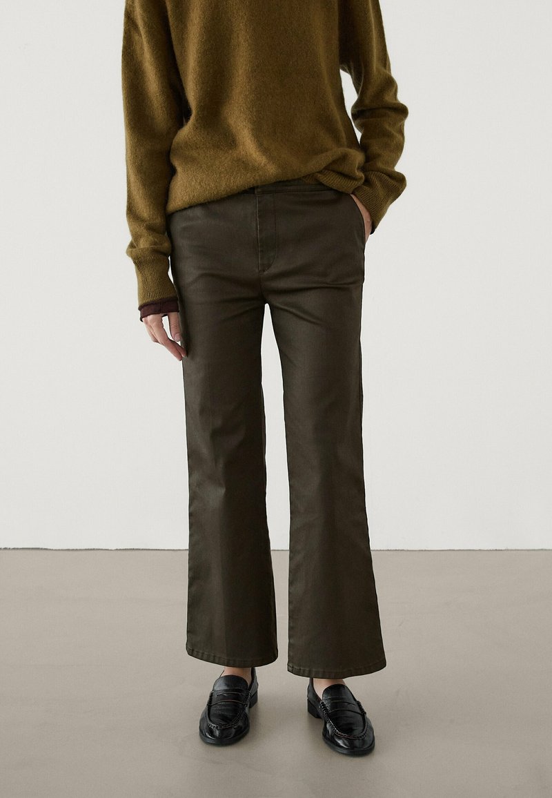 Personne portant un pantalon ample vert olive, un pull marron et des mocassins noirs, debout sur un sol gris devant un mur blanc.