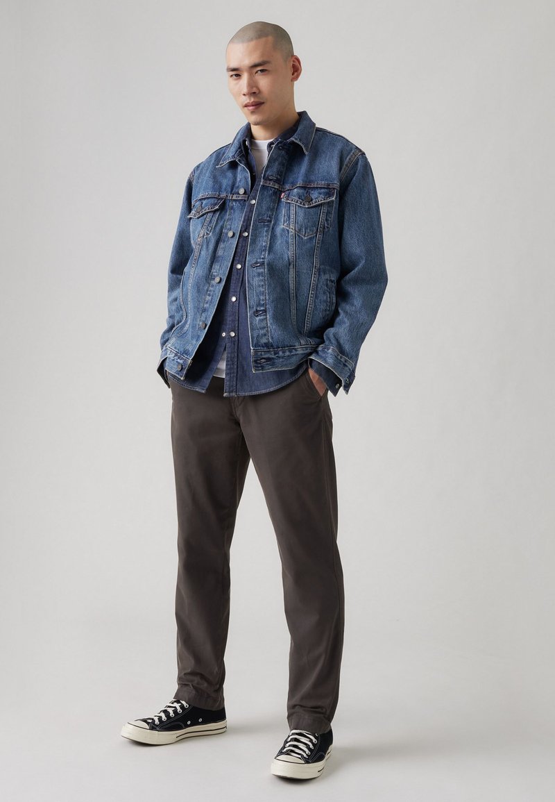 Chaqueta de mezclilla en azul medio con dos bolsillos en el pecho, cierre de botones y un cuello clásico. Combinada con pantalones oscuros grises y zapatillas negras.