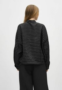 Fekete és szürke kockás flaneling, laza szabással, lekerekített gallérral, hosszú ujjal, és egy diszkrét zseb részlettel a hátán.