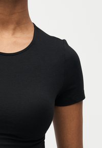 Κοντινό πλάνο προσώπου που φοράει εφαρμοστό μαύρο κοντομάνικο μπλουζάκι με στρογγυλή λαιμόκοψη σε λευκό απλό φόντο.