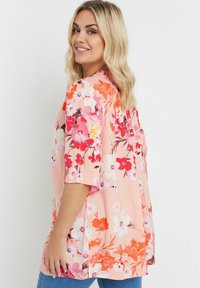 Blusa floreale in morbido tessuto rosa, caratterizzata da maniche corte, fiori arancioni e rossi audaci, e un fit rilassato con spacchi laterali.