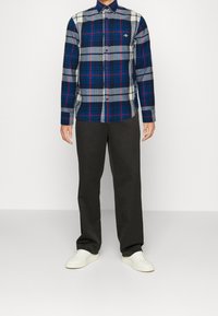 Camisa de manga larga a cuadros en azul, gris y blanco con cuello abotonado. Combinada con pantalones oscuros y zapatillas blancas.