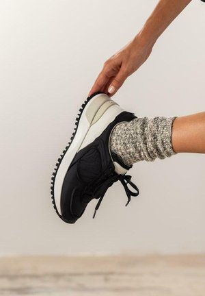 Zapatilla deportiva negra con una parte superior texturizada, media suela blanca y suela negra; combinada con un calcetín gris de punto.