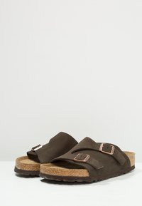 Brown suede slide sandals with two adjustable straps, cork footbed, and rubber sole featuring textured tread for grip.