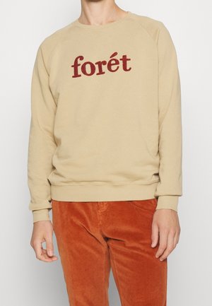 Person iført beige sweatshirt med rød "forét" tekst og rustfarvede manchesterbukser, stående mod en ensfarvet lys baggrund.