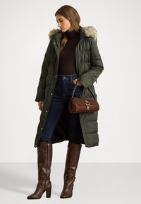Olívovo zelená prešívaná bunda s umelou kožušinou na kapucni, priliehavý strih, kombinovaná s tmavými džínsami a hnedými vysokými čižmami po kolená, drží hnedú semišovú kabelku.