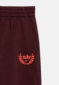 Burgundijas krāsas Nike sporta bikses ar elastīgu jostu un oranžu izšūtu logo, kas satur lauru dizainu apakšējajā kreisajā stūrī. Mīksta tekstūra.