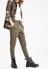 Olivgröna smalna byxor med elastisk midja och snörning, tillsammans med en vit skjorta och svarta snörboots i combat-stil. Camo-jacka som bärs öppen.