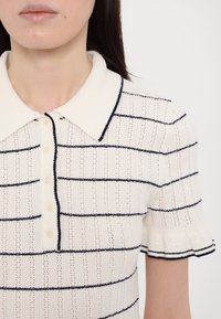 Polo en tricot rayé couleur crème, avec des rayures horizontales bleues marines. Texture douce, fermeture à boutons et manches courtes à volants.