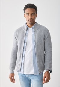 Grå ribbestrikket cardigan med glidelås foran, båret over en lyseblå skjorte med knapper, kombinert med en hvit T-skjorte og lyseblå jeans.