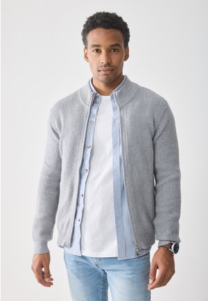 Graue gerippte Strickjacke mit Reißverschluss, getragen über einem hellblauen Button-up-Hemd, kombiniert mit einem weißen T-Shirt und hellblauen Jeans.